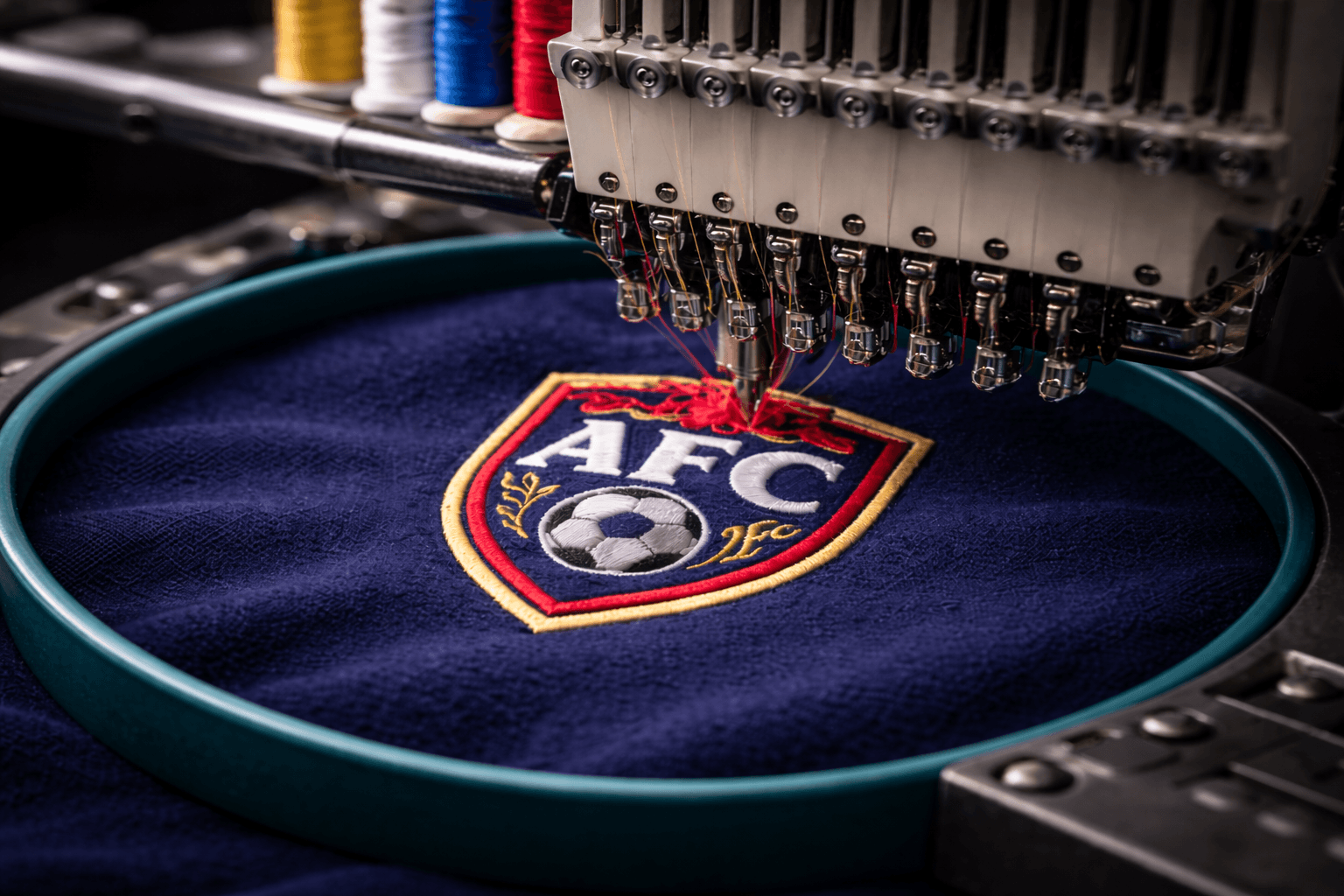 Industrial embroidery machine stitching a colorful AFC soccer crest onto dark blue fabric material.