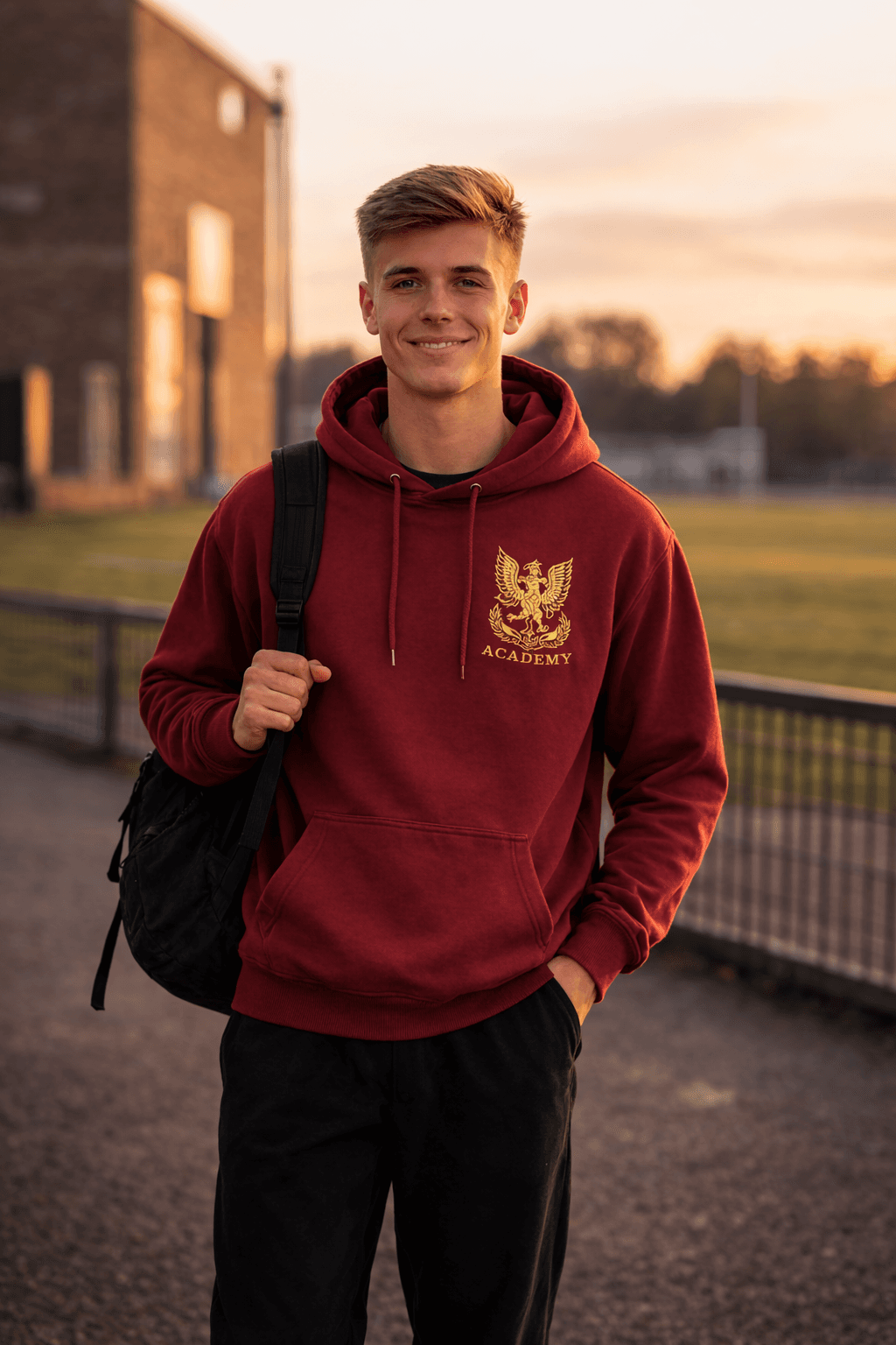 Smiling young man wearing a maroon academy hoodie and backpack on campus at sunset.