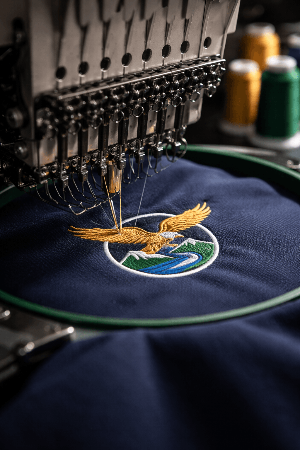 Industrial embroidery machine stitching a golden eagle and mountain logo onto dark blue fabric.